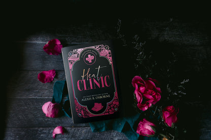 An aesthetic photo of the Heat Clinic special edition hardcover with the discreet dust jacket showing pink images on a black background. There are pink flowers and a teal ribbon surrounding the book on a dark wooden floor.